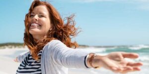 Beautiful middle aged woman with red hair and arms up dancing on beach in summer during holiday, meditation-changes-your-appearance