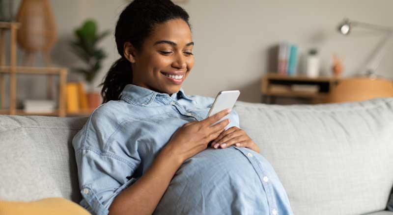 A smiling pregnant woman sitting on a couch looking at her phone