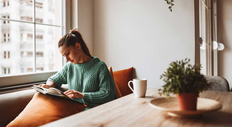 A woman sitting indoors by a window reading a book