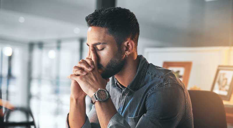 A man sitting at a desk with his eyes closed and head in his hands